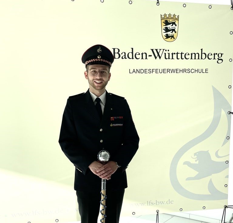 Matthias Molnar steht in Uniform mit Stab vor einem Plakat mit der Aufschrift "Baden-Württemberg - Landesfeuerwehrschule" und lächelt in die Kamera