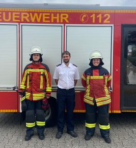 Die beiden neuen Truppführer*innen stehen in Einsatzuniform vor einem Feuerwehrfahrzeug, zwischen ihnen Abteilungskommandant Matthias Erck